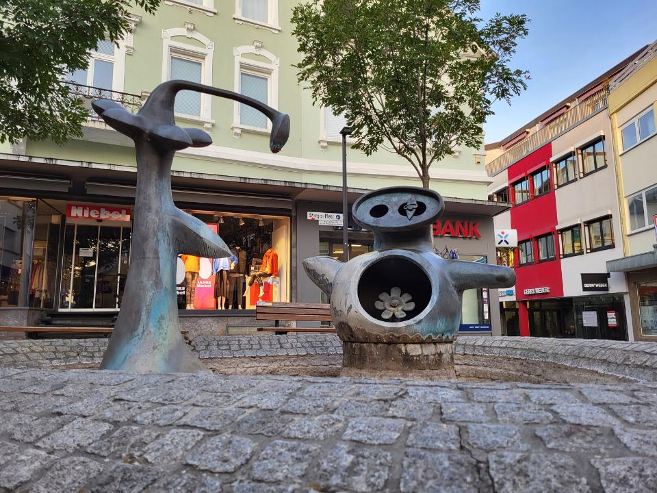 2 Bronze-Figuren die an Blumen erinnern. Die Blumen können Wasser speien, wenn der Brunnen angeschaltet ist.