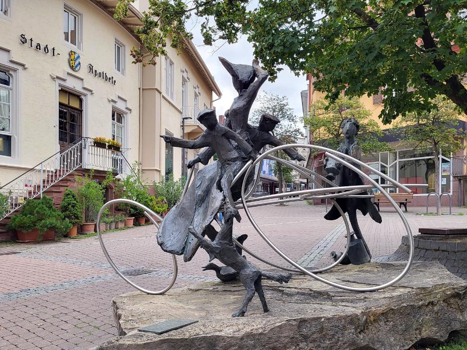 Abstrakte Skulptur aus Bronze und Edelstahl, 1 Frau und 2 Kinder sitzen auf einem Patent-Motorwagen.